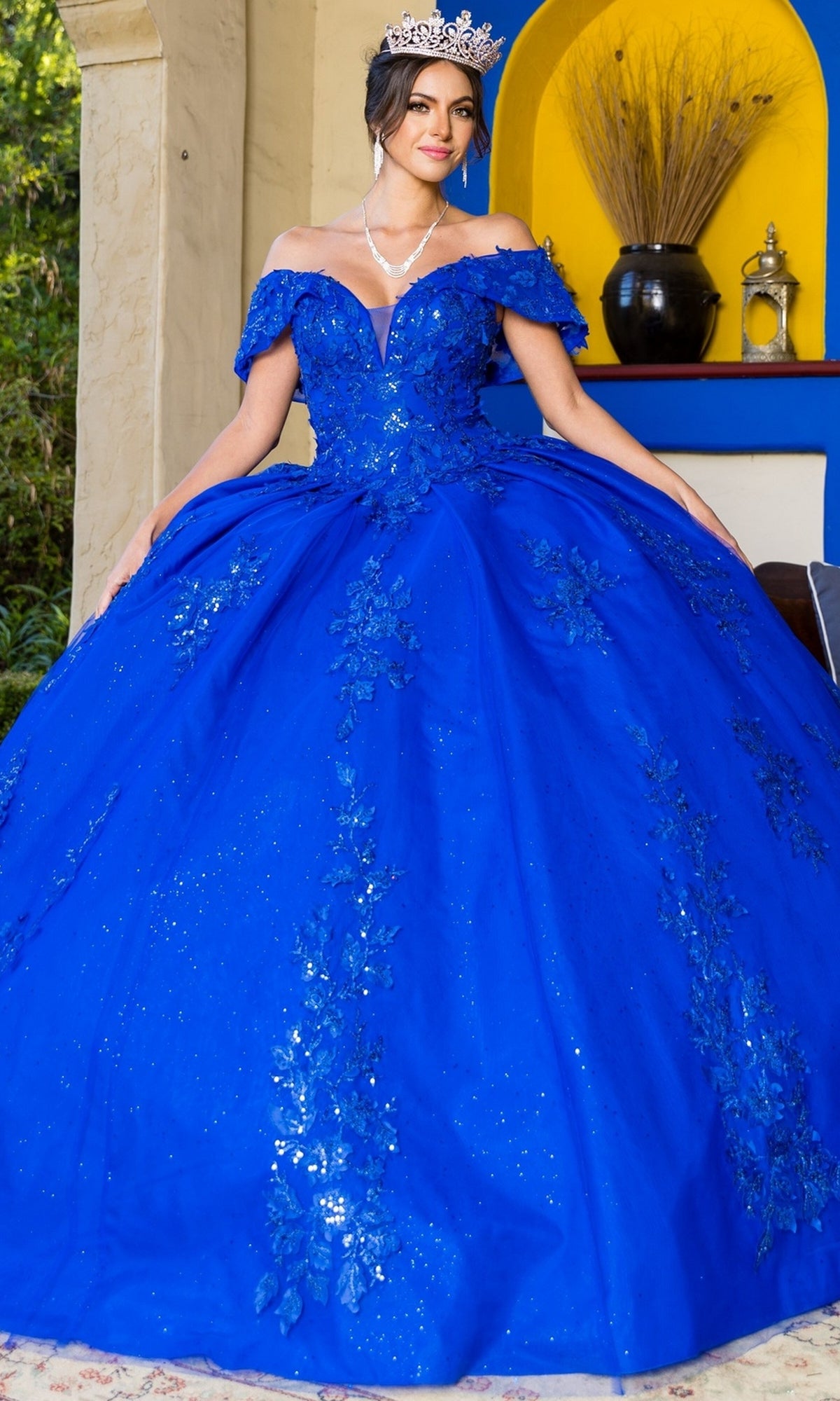 A person in the Quinceanera Ball Gown 8045J, featuring ornate 3D floral details and an off-the-shoulder royal blue design with a silver crown, stands before a colorful alcove.